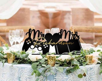 Op de afbeelding: Een bruidstaarttopper met een zwart silhouet van een videogamecontroller en de woorden "Mr. & Mrs. Richardson" in gouden letters.