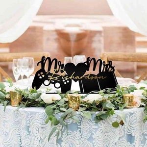 May include: A wedding cake topper with a black silhouette of a video game controller and the words "Mr. & Mrs. Richardson" in gold lettering.