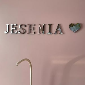 May include: A pink wall with the name "JESENIA" in silver letters and a silver heart with a floral design.
