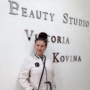 May include: A woman with dark hair pulled back in a bun stands in front of a white wall with the words "Beauty Studio Victoria Kovina" written in silver letters.