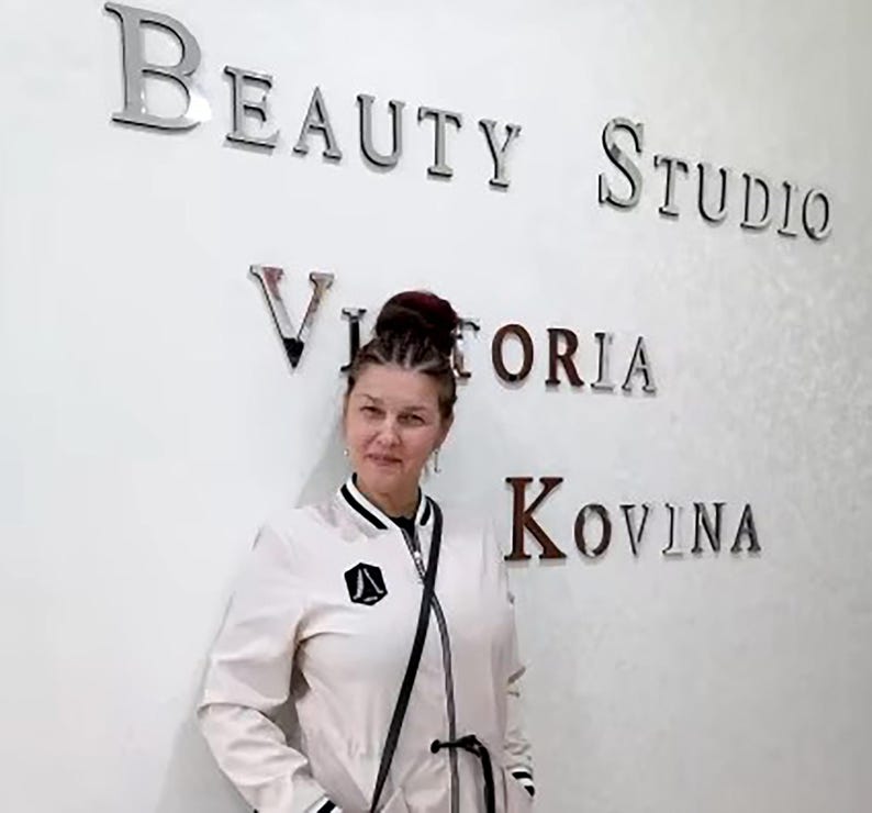May include: A woman with dark hair pulled back in a bun stands in front of a white wall with the words "Beauty Studio Victoria Kovina" written in silver letters.