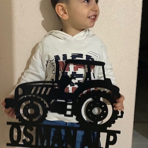 May include: A black metal tractor cutout with the name "OSMAN ALP" underneath. A young child is holding the cutout in front of them.