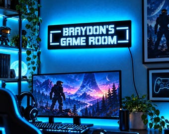 May include: A rectangular wall sign with a black background and bright blue LED backlighting, featuring the white text "BRAYDON'S GAME ROOM" in a blocky, digital-style font, mounted on a wall above a computer monitor and desk setup.