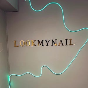 May include: A gold sign with the text "LOOKMYNAIL" on a white wall with a blue neon light in the shape of a wave.