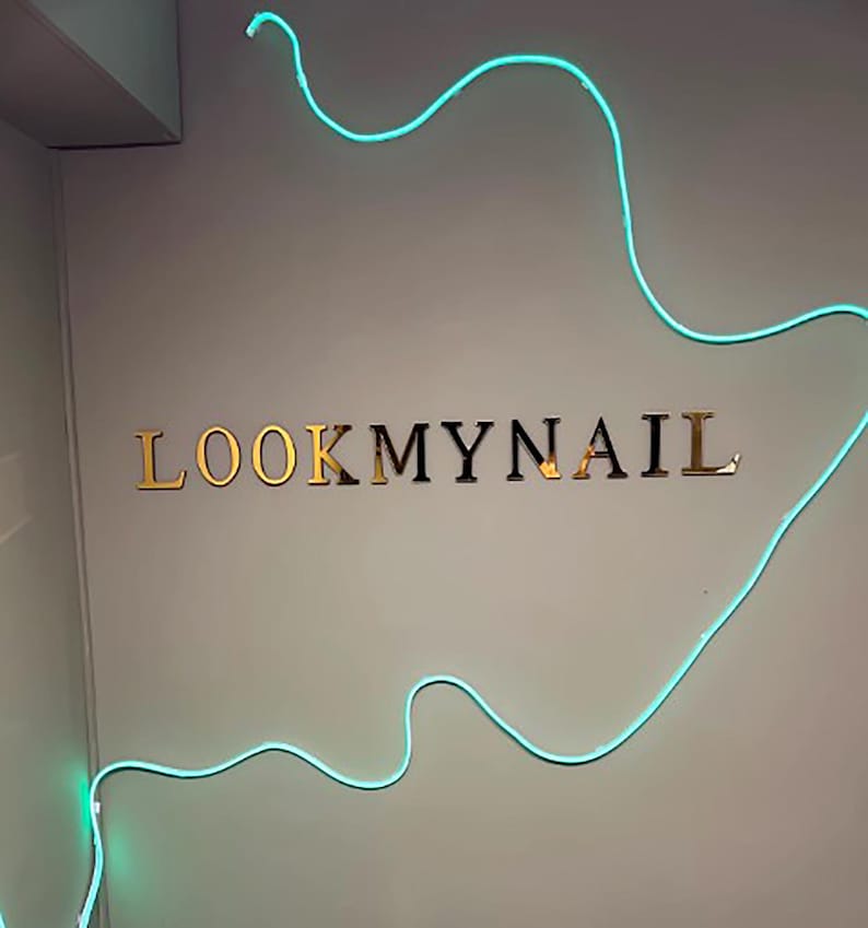 May include: A gold sign with the text "LOOKMYNAIL" on a white wall with a blue neon light in the shape of a wave.