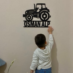 May include: Black wooden tractor wall decor with the name "OSMAN ALP" underneath. A young child is reaching up to touch the decor.