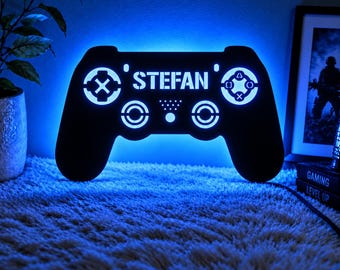 May include: A black wall-mounted gaming controller sign featuring a bright blue LED backlight. The center of the controller displays the name STEFAN in bold white letters, surrounded by circular button icons and a small dot pattern.