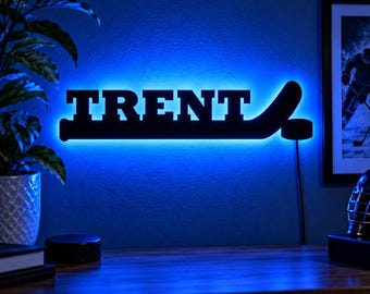 May include: A black wall-mounted sign featuring the name TRENT in bold capital letters integrated into a hockey stick shape, illuminated from behind with a vibrant blue LED light that casts a glow against the wall surface.