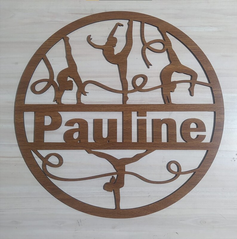 May include: A brown wooden circle with the name "Pauline" cut out in the center. The circle features a silhouette of four gymnasts performing on a ribbon.