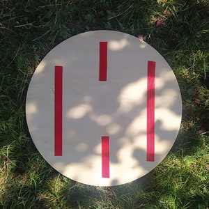 May include: A round wooden disc with four red stripes painted on it. The disc is sitting on a bed of green grass.
