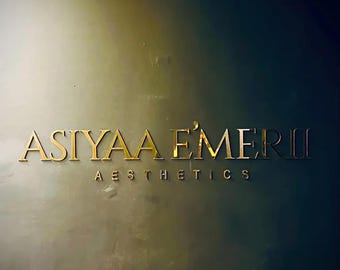 May include: Gold metallic sign with the name "ASIYAA EMERII" and the word "AESTHETICS" below it. The sign is mounted on a wall with a gradient of colors, transitioning from a light gold to a dark blue.
