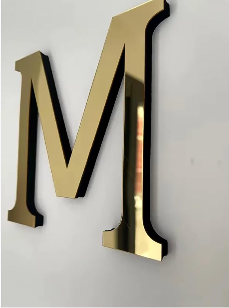 May include: A gold-colored letter M with a black border. The letter is made of a reflective material, and the surface is smooth.