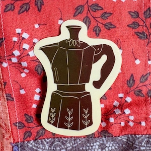 May include: A brown sticker featuring a coffee pot design. The sticker is placed on a patchwork background of red floral fabric with white accents, and other patterned fabrics. The coffee pot has a handle and decorative leaf details.