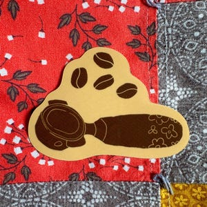 May include: A tan sticker featuring a coffee portafilter and coffee beans. The sticker is set against a patchwork background of red floral fabric, gray paisley fabric, and yellow floral fabric.