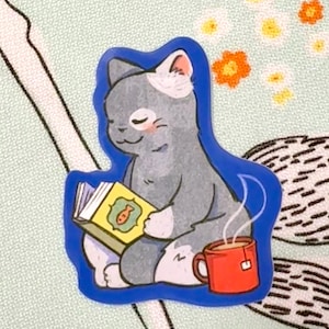 May include: A sticker featuring a gray cat with white markings, reading a book with a fish illustration. The cat is sitting next to a red mug of tea. The sticker has a blue outline.