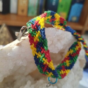 Op de afbeelding: Een kleurrijk vriendschapsarmband met rode, gele, blauwe en groene draden die samen in een chevron-patroon zijn geweven. De armband heeft een zilveren bedel eraan en de woorden "Opale Rose" staan op het oppervlak onder de armband.