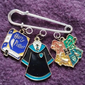 Peut inclure: Une épingle de sûreté argentée avec trois breloques émaillées. Les breloques comprennent un livre bleu et or avec le texte "Harry Potter", une robe noire et sarcelle et un blason avec des sections rouges, or, vertes et bleues.