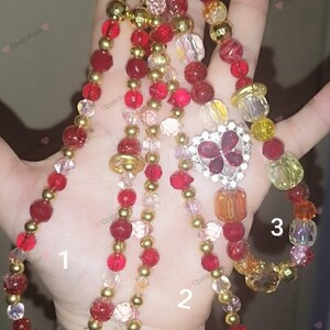 Op de afbeelding: Drie kettingen met kralen in rood, goud en helder. Eén ketting heeft een hartvormige hanger met rode stenen. De kettingen variëren in lengte en kraalgrootte, met goudkleurige accenten.