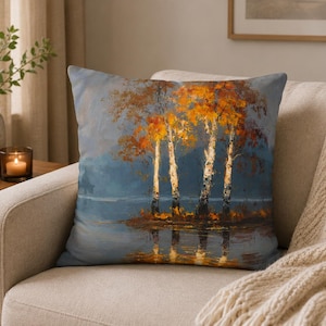 Könnte beinhalten: Ein quadratisches Zierkissen mit einem Ölgemälde-Druck, der eine Gruppe von Birken mit leuchtend orangefarbenen und gelben Herbstblättern zeigt. Die Bäume sind vor einem gedämpften blauen Hintergrund dargestellt, ihre Spiegelung ist im Wasser sichtbar.
