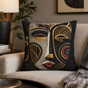 Könnte beinhalten: Schwarzes Zierkissen mit einem abstrakten Gesichtsdesign in Gold, Rot und Grau. Das quadratische Kissen liegt auf einem beigen Sofa und verleiht dem Raum eine künstlerische Note.