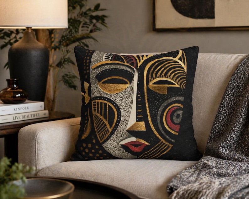May include: Black decorative throw pillow featuring an abstract face design with gold, red, and gray accents. The pillow is square-shaped and rests on a beige sofa, adding a touch of artistic flair to the living space.