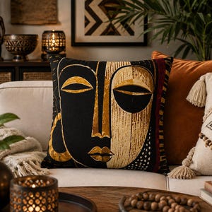 May include: A square throw pillow featuring a stylized face design split into black and gold sections. The left side is solid black with a gold earring, while the right side displays a textured gold pattern. The design includes almond-shaped eyes and defined lips, with a dark red and gold striped border on the right edge.