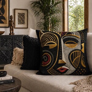 May include: Decorative throw pillows with an African mask design. The pillows feature a black background with gold, red, and white accents. The pillows are on a beige sofa with a woven throw blanket.