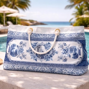 Könnte beinhalten: Eine große weiße und blaue Tragetasche mit Blumen- und Landschaftsdesign. Die Tasche hat einen Seilgriff und goldfarbene Ringe. Ein Strohhut mit schwarzem Band liegt vor der Tasche, in der Nähe eines Pools.