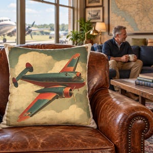 Könnte beinhalten: Ein quadratisches Zierkissen mit einer Illustration im Vintage-Stil eines zweimotorigen Propellerflugzeugs im Flug. Das Flugzeug ist in Blaugrün und Rot gehalten und vor einem Hintergrund aus cremefarbenen Wolken mit dezentem Kartenmuster abgebildet.