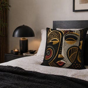 May include: Decorative throw pillow with a black background and a stylized face design in gold, red, and white. The pillow is placed on a bed with a black blanket and pillows. A black lamp and a candle are on the nightstand.