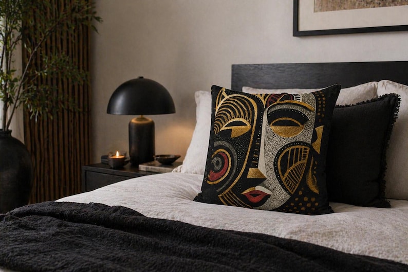 May include: Decorative throw pillow with a black background and a stylized face design in gold, red, and white. The pillow is placed on a bed with a black blanket and pillows. A black lamp and a candle are on the nightstand.