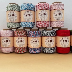 May include: A collection of twelve spools of baker's twine in various colors, including green, blue, red, and pink. Each spool has a brown label with the text "Compostable Bakers Twine" and a scissors graphic. The twine is arranged in neat rows.