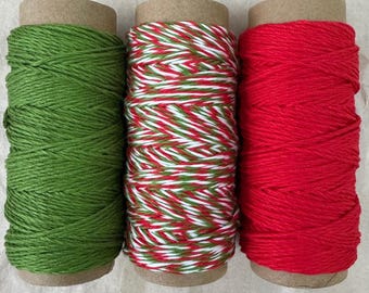 May include: Three spools of twine in festive colors. One is solid green, the second is a mix of red, green, and white, and the third is solid red. Each spool is wrapped around a cardboard tube, perfect for crafting or holiday projects.