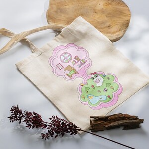 Pu&ograve; includere: Borsa tote in tela beige con illustrazioni di cartoni animati rosa e verdi. Un'illustrazione raffigura una stanza con finestra, sedia e libri. L'altra mostra un animale in un parco.