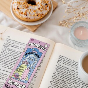 Puede incluir: Un marcap&aacute;ginas rosa y azul con una luz adjunta. El marcap&aacute;ginas tiene las palabras "Midnight Reading Light" y "Reader Essentials" en &eacute;l. El marcap&aacute;ginas est&aacute; sobre un libro con una taza de caf&eacute; y una vela en el fondo.