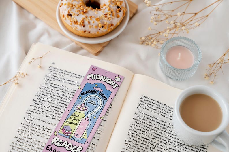 Puede incluir: Un marcap&aacute;ginas rosa y azul con una luz adjunta. El marcap&aacute;ginas tiene las palabras "Midnight Reading Light" y "Reader Essentials" en &eacute;l. El marcap&aacute;ginas est&aacute; sobre un libro con una taza de caf&eacute; y una vela en el fondo.