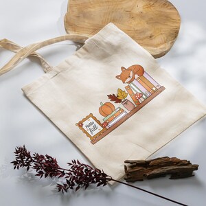 以下が含まれることがあります： 本の上に寝ているキツネ、カボチャ、そして「Hello Fall」の文字が描かれた、カラフルなイラストが特徴のベージュのトートバッグ。デザインには秋の葉やキノコが含まれています。バッグには2つのベージュのハンドルが付いています。