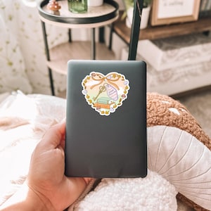 May include: A black laptop with a heart-shaped sticker. The sticker features a key, books, and a bottle with the text "Dream Follower" on a ribbon. The sticker is surrounded by a floral design with a light orange and yellow color scheme.