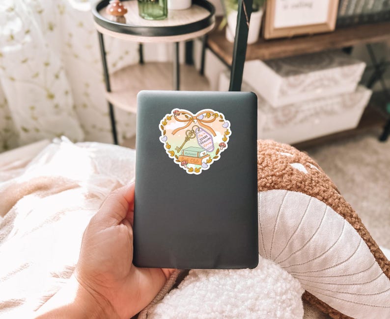 May include: A black laptop with a heart-shaped sticker. The sticker features a key, books, and a bottle with the text "Dream Follower" on a ribbon. The sticker is surrounded by a floral design with a light orange and yellow color scheme.