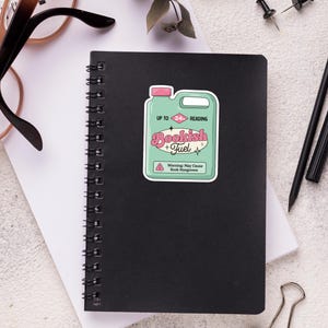 May include: A green and pink sticker with a cartoon illustration of a fuel container labeled "Bookish Fuel" with the text "Up to 24 Hours Reading" and a warning that it may cause "Book Hangovers".