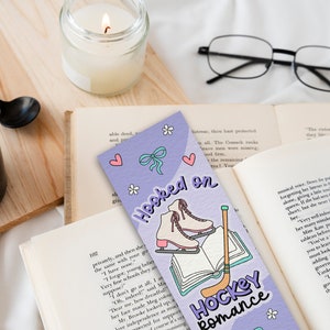 May include: Purple bookmark with a light blue bow, white ice skates, a book, and a hockey stick. The bookmark has the text "Hooked on Hockey Romance".