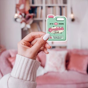 May include: Green and pink sticker with the text "Bookish Fuel" and "Up to 24+ Reading" in a retro font. The sticker is shaped like a jug of liquid with a warning label that reads "Warning: May Cause Book Hangovers".