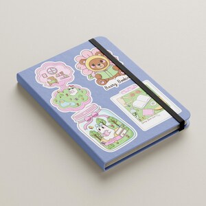 Pu&ograve; includere: Un quaderno azzurro con una fascia elastica nera. &Egrave; decorato con adesivi colorati raffiguranti orsetti dei cartoni animati, fiori e altri disegni stravaganti. Il testo "Beary Bookin" &egrave; visibile su uno degli adesivi.