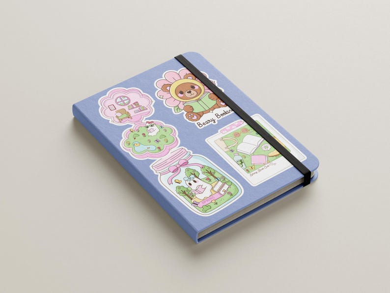 Pu&ograve; includere: Un quaderno azzurro con una fascia elastica nera. &Egrave; decorato con adesivi colorati raffiguranti orsetti dei cartoni animati, fiori e altri disegni stravaganti. Il testo "Beary Bookin" &egrave; visibile su uno degli adesivi.