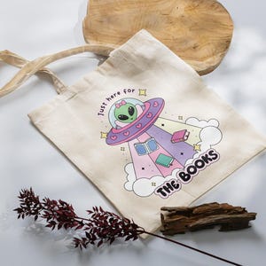 May include: A white canvas tote bag with a graphic of a green alien in a pink and purple spaceship with the text "Just here for the books".