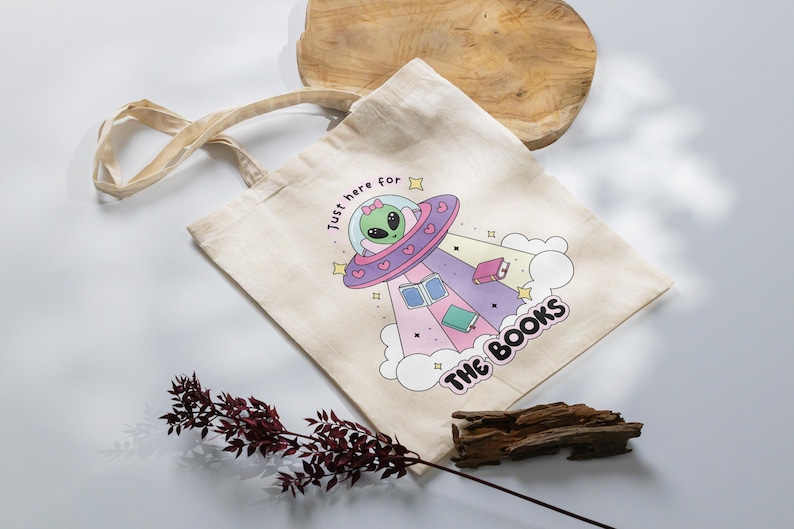 May include: A white canvas tote bag with a graphic of a green alien in a pink and purple spaceship with the text "Just here for the books".