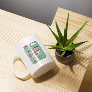 May include: White ceramic mug with a green and pink design of a bottle labeled "Beach Fuel". The mug is sitting on a wooden surface with a green plant in a gray pot.
