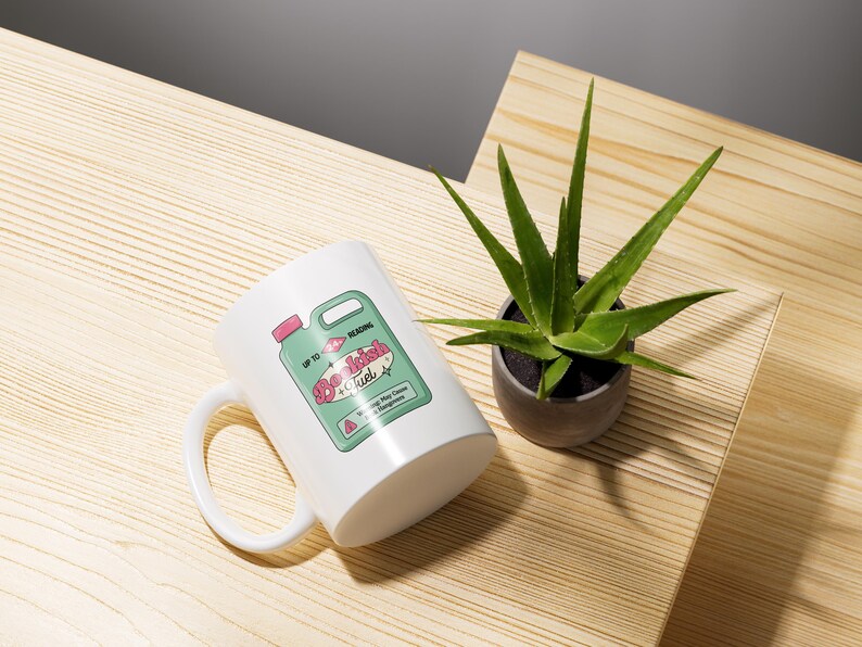 May include: White ceramic mug with a green and pink design of a bottle labeled "Beach Fuel". The mug is sitting on a wooden surface with a green plant in a gray pot.