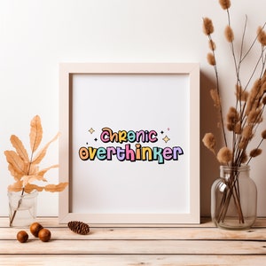 May include: A white framed print with the text "Chronic overthinker" in colorful letters with a rainbow color scheme. The print is on a white background with small stars around the text. The print is sitting on a wooden table with a vase of dried flowers and a few acorns.