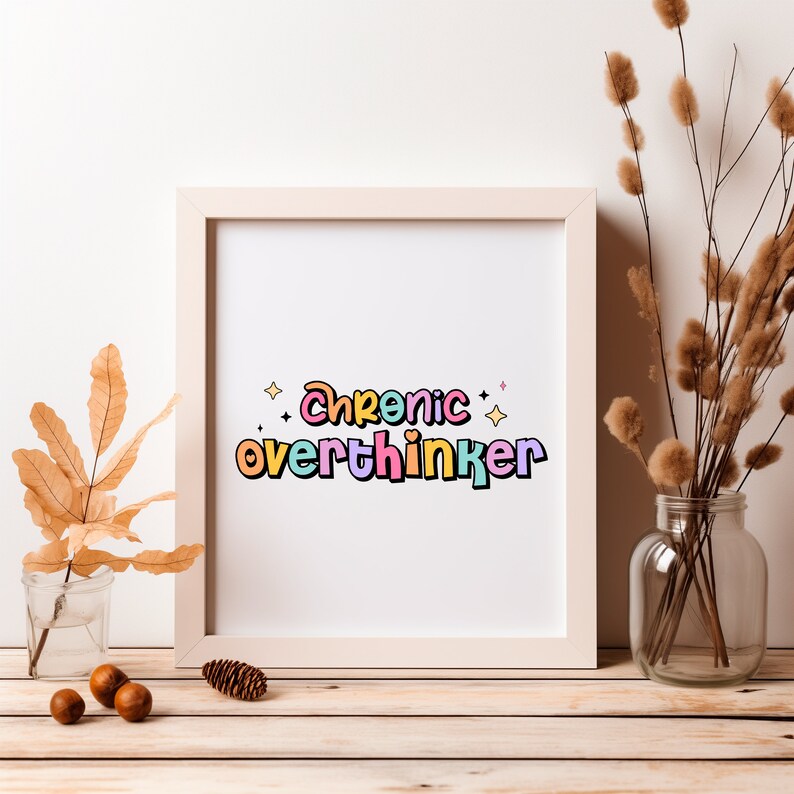 May include: A white framed print with the text "Chronic overthinker" in colorful letters with a rainbow color scheme. The print is on a white background with small stars around the text. The print is sitting on a wooden table with a vase of dried flowers and a few acorns.
